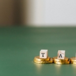 Stacked gold coins with wooden blocks spelling 'TAX' on a green backdrop.