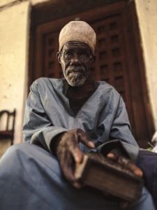 An elderly man in traditional attire reads outdoors in Tanzania, capturing cultural depth and wisdom.