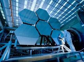 Engineers in protective suits work on telescopic mirrors in a high-tech lab.