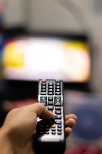 Close-up of a hand holding a remote control, watching TV indoors. Captures modern leisure and technology.