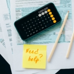 Top view of financial papers with a calculator, pencils, and a note saying 'Need help?' indicating tax or accounting assistance.