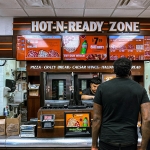 Busy fast food restaurant with customers ordering pizza at the counter under bright digital menu signage.